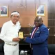 Menteri HAM Natalius Pigai dalam balutan jas biru bertemu dengan Gubernur Jawa Barat Dedi Mulyadi yang mengenakan pakaian putih dan ikat kepala, keduanya bertukar cendera mata di ruang pertemuan resmi dengan lambang Garuda Pancasila dan foto Presiden sebagai latar belakang.