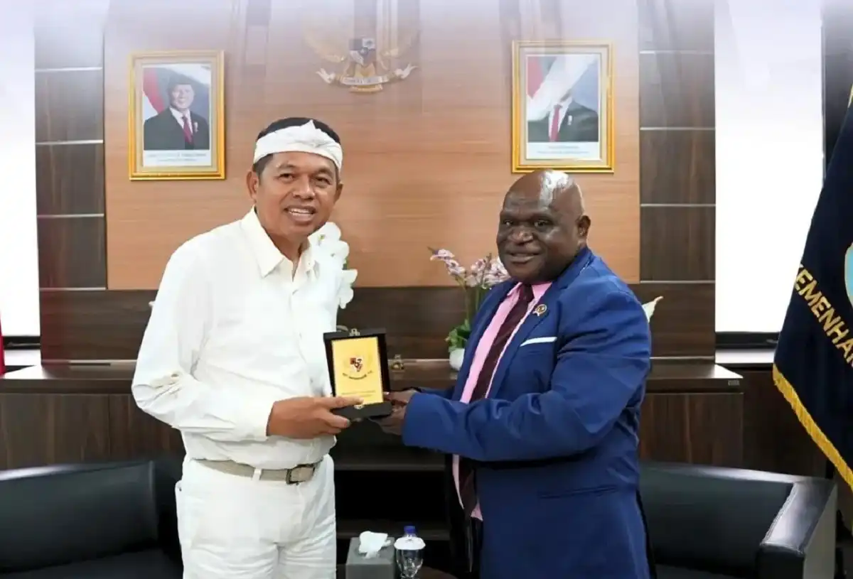Menteri HAM Natalius Pigai dalam balutan jas biru bertemu dengan Gubernur Jawa Barat Dedi Mulyadi yang mengenakan pakaian putih dan ikat kepala, keduanya bertukar cendera mata di ruang pertemuan resmi dengan lambang Garuda Pancasila dan foto Presiden sebagai latar belakang.
