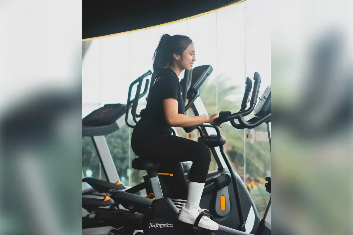 Seorang wanita tersenyum bahagia saat menggunakan elliptical trainer di gymnasium, mencerminkan filosofi mengolah raga dan jiwa untuk refleksi diri di tengah padatnya rutinitas Jabodetabek