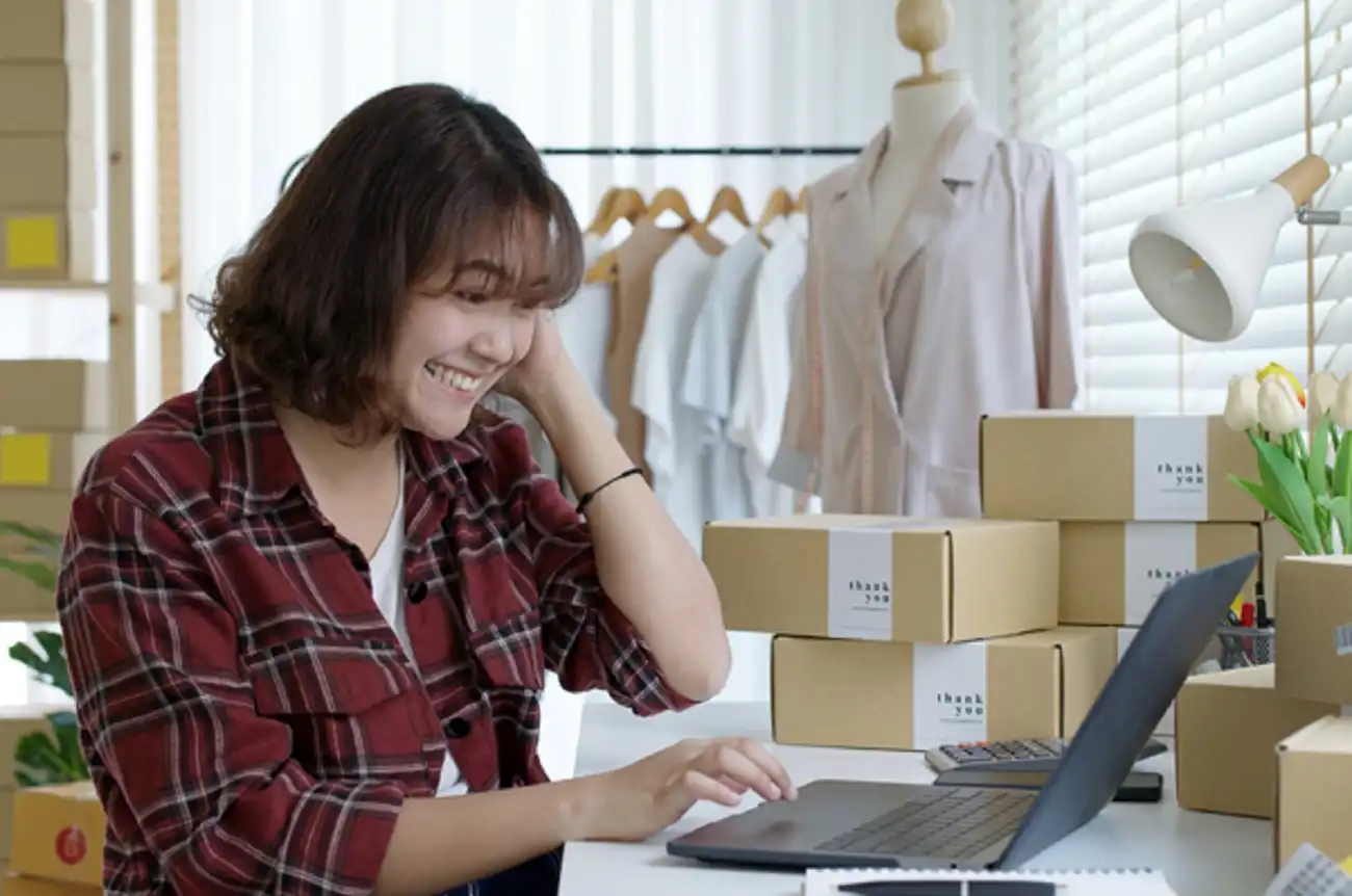 Seorang pelaku UMKM sedang mengelola penjualan online di depan laptop, dikelilingi paket produk siap kirim.